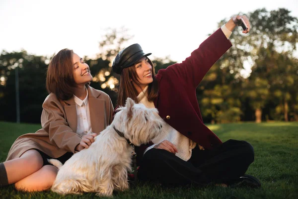 Porträtt av två unga leende damer sitter på gräset och tar selfie på mobiltelefon samtidigt spendera tid med sin lilla söta hund i parken — Stockfoto