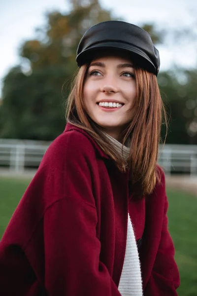 Portrait of beautiful lady in black cap and coat standing in park and happily looking aside isolated — Stock Photo, Image