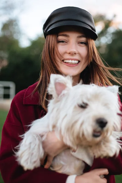 Porträtt av glada vackra dam i svart keps och pälsen står i park och glatt tittar i kameran medan du håller i händerna hennes små söta hund — Stockfoto