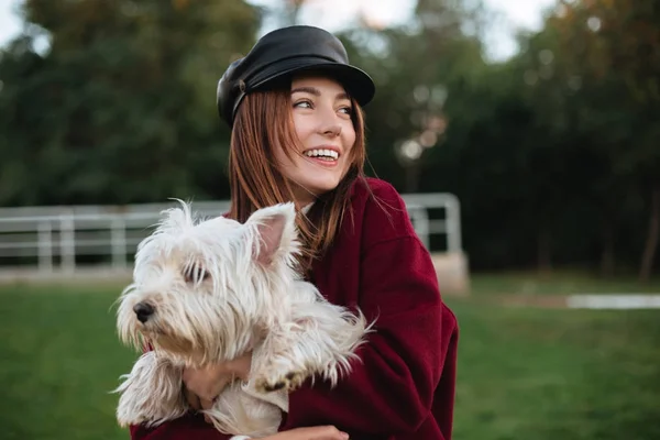 戴着黑色帽子和大衣的微笑美丽的女士的肖像站在公园, 并高兴地看着一旁, 而手牵着她的小可爱的狗 — 图库照片