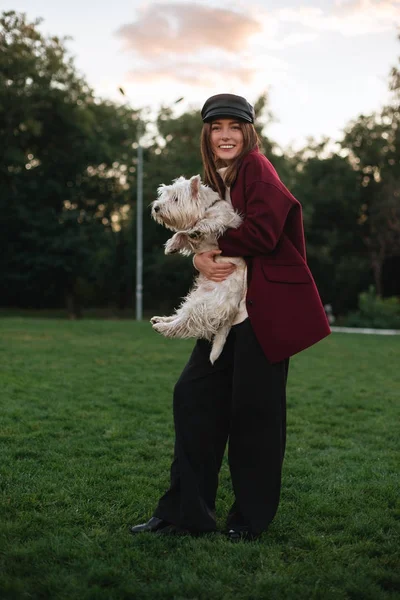 Bella signora sorridente in berretto nero e cappotto in piedi nel parco e gioiosamente guardando in macchina fotografica mentre abbraccia il suo piccolo cane carino — Foto Stock