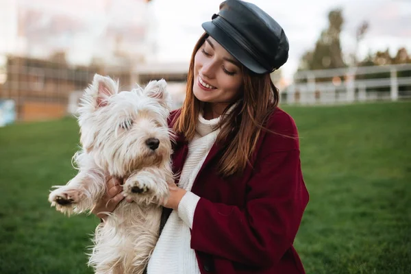Porträtt av ung vacker dam i svart keps och pälsen håller i händer hennes små söta hund medan spendera tid i park — Stockfoto