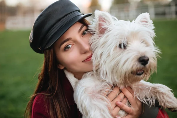 Ritratto di giovane bella signora in berretto nero e cappotto in piedi e sognante guardando in macchina fotografica mentre abbraccia il suo piccolo cane carino nel parco — Foto Stock