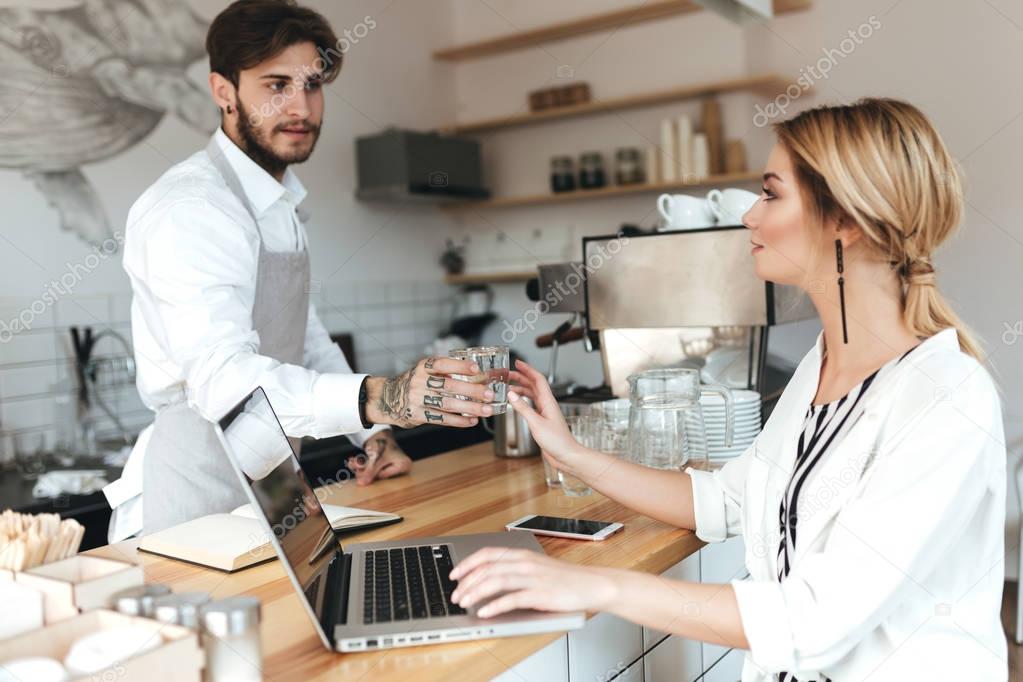 Young barista in apron and white shirt giving glass of water to pretty girl. Beautiful lady with blond hair sitting at the counter and working on her laptop in coffee shop