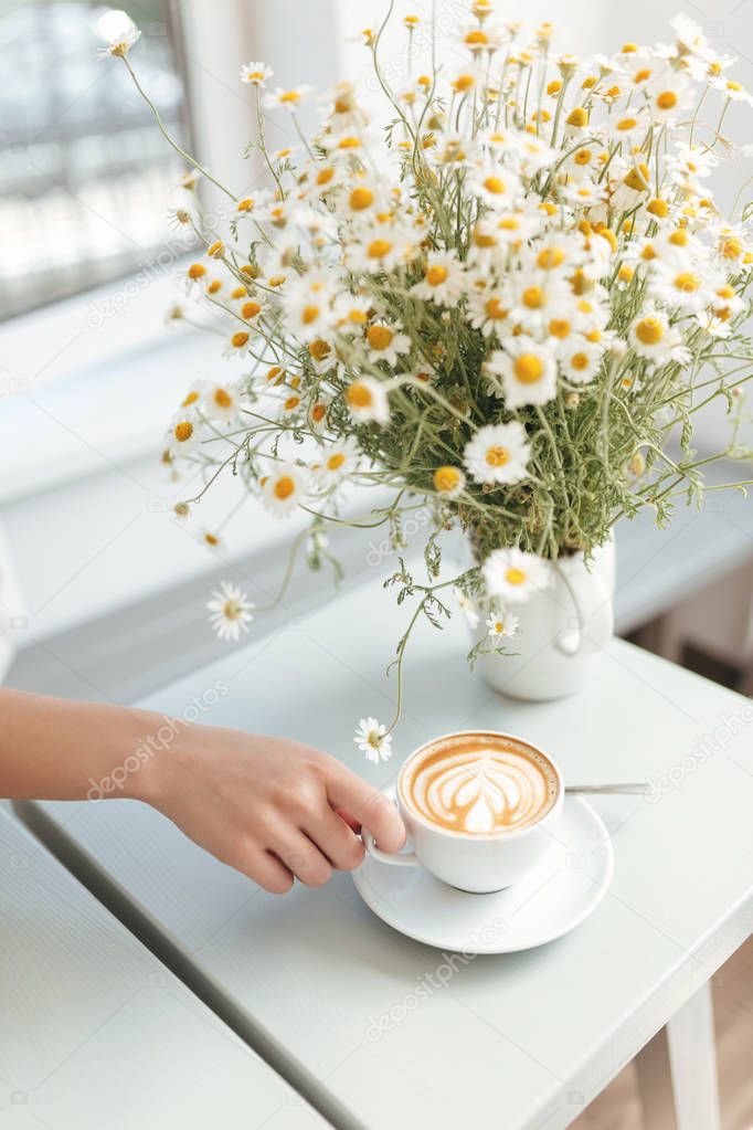 Beautiful close up photo of chamomile flowers and white cup of cappuccino on table in coffee shop. Woman hand holding cup of coffee in restaurant