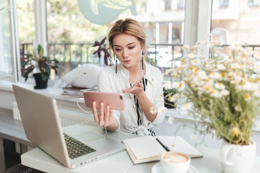 Portrait of young girl in earphones looking in her mobile phone and sending kisses in coffee shop. Nice girl with blond hair sitting in restaurant with laptop,notebook and cup of coffee on table