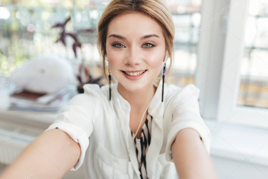 Beautiful young girl making photos on her frontal camera in mobile phone. Pretty smiling lady with blond hair in earphones happily looking in camera in restaurant. Cool girl wearing white shirt