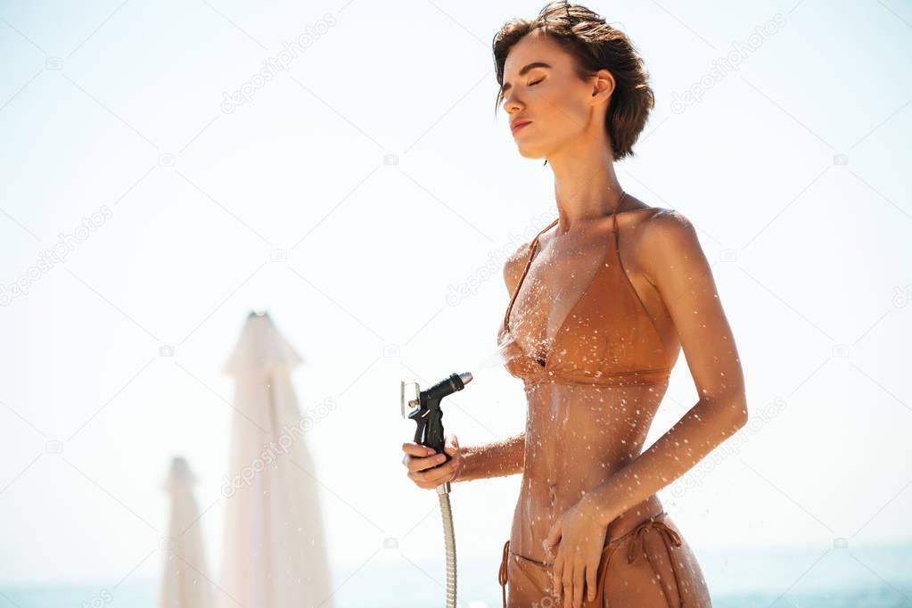 Portrait of girl in bikini using hose pipe on beach. Young lady in beige swimsuit rinsing beach sand off her body on beach. Pretty girl dreamily closing her eyes while pouring water from hose on beach