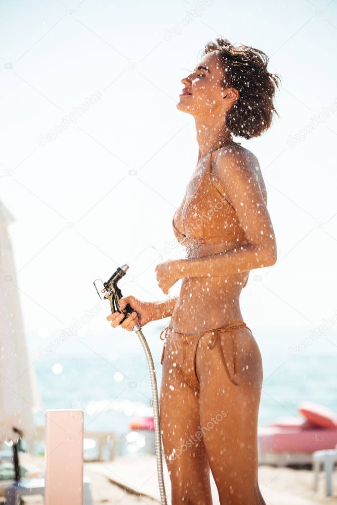 Portrait of cute girl in bikini standing on beach and using hose pipe. Young lady in beige swimsuit rinsing beach sand off her body on beach. Nice girl pouring water from hose while standing on beach