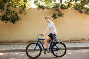 Sersem kamera bisiklet sürme sırasında arıyorum boy gülümseyerek portresi. Şehrin caddeleri boyunca mavi bisiklet sürme sırasında eğlenmek sarışın saçlı genç adam