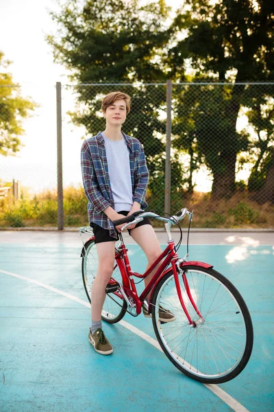 Młody człowiek z blond włosami w szorty i casual shirt stojący z czerwony rower na boisko do koszykówki w parku. Portret chłopca, starannie patrząc w aparacie stojąc z klasyczny rower — Zdjęcie stockowe