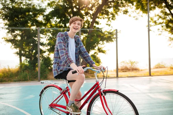 Portret wesoły chłopak o blond włosach w szorty i casual shirt jazdy na rowerze na boisko do koszykówki w parku. Uśmiechnięty chłopiec szczęśliwie patrząc na bok stojąc z rowerem — Zdjęcie stockowe