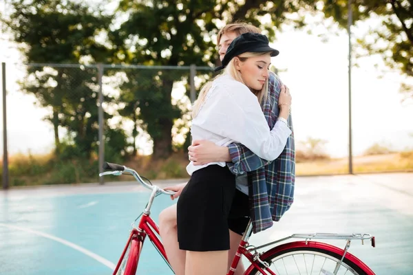 Young pretty girl with blond hair dreamily closing her eyes while embracing boy on red bicycle on basketball court in park. Portrait of beautiful couple embracing one another — Stock Photo, Image