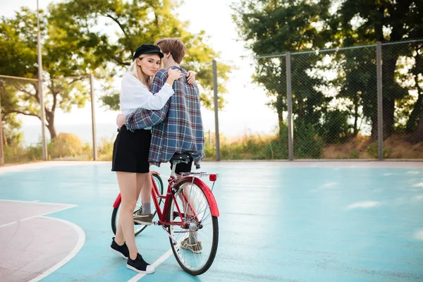 Hermosa chica con el pelo rubio cuidadosamente mirando en la cámara mientras abraza niño en bicicleta roja clásica en la cancha de baloncesto en el parque. Retrato de una linda pareja joven abrazándose —  Fotos de Stock