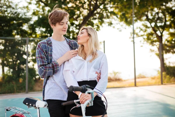 Menina bonita com cabelo loiro e menino sonhadoramente olhando um para o outro, enquanto em pé no parque. retrato de bonito jovem casal abraçando um ao outro com bicicleta ao lado — Fotografia de Stock