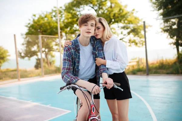 Niño en bicicleta soñando mirando en la cámara mientras hermosa chica con el pelo rubio de pie al lado y abrazándolo en el parque. Retrato de linda pareja joven con bicicleta en cancha de baloncesto — Foto de Stock