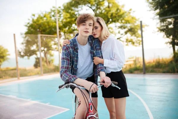 Jongen op fiets dromerig op zoek in de camera terwijl mooi meisje met blond haar permanent naast en hem omhelzen in park. Portret van schattig jong koppel met fiets op het basketbalveld — Stockfoto