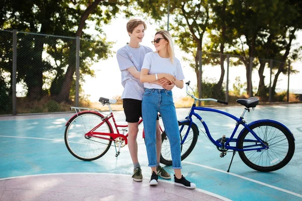 Retrato de pareja joven felizmente mirándose unos a otros con bicicletas rojas y azules en el fondo. Alegre chico de pie con hermosa chica en gafas de sol y abrazándola mientras pasa tiempo en el parque —  Fotos de Stock