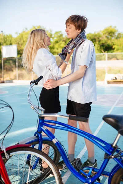 男孩和女孩与金发碧眼的花时间在一起, 并在公园的篮球场乐趣。年轻夫妇的肖像站在一起玩蓝色和红色的自行车旁边 — 图库照片