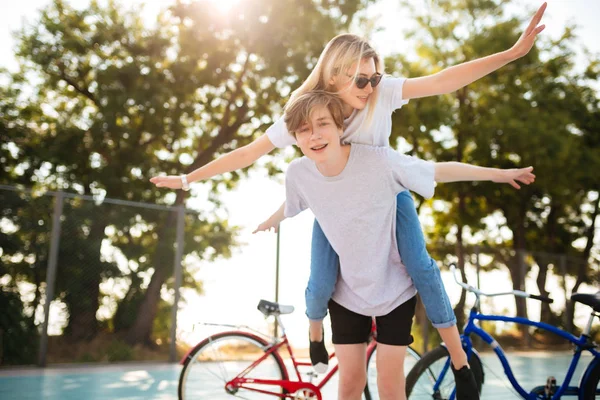 Porträtt av unga par att ha roligt tillsammans i parken med cyklar på bakgrunden. Leende pojke leker med vacker flicka i solglasögon och visar plan vingar gest medan spendera tid i park — Stockfoto