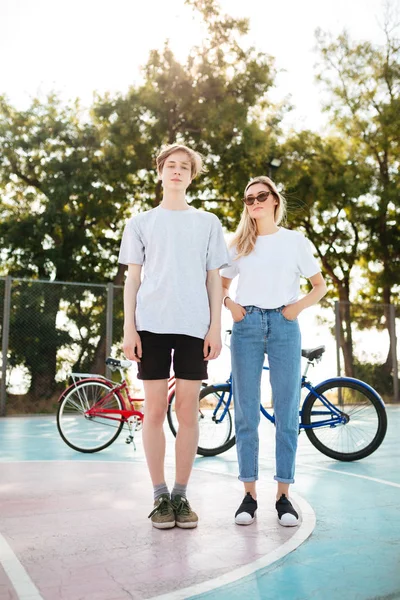 Söt tjej med blont hår i solglasögon och cool kille står och eftertänksamt tittar i kameran med två cyklar på bakgrund. Unga par tillbringa tid tillsammans i basketplan — Stockfoto