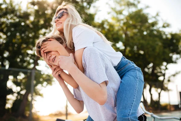 Портрет молодой пары, развлекающейся вместе в парке. Веселый мальчик играет с красивой девушкой в солнечных очках и держит ее на спине, пока она игриво закрывает ему глаза — стоковое фото