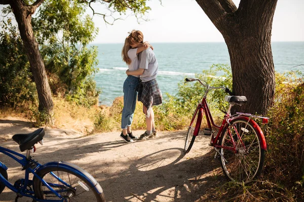 Retrato de una hermosa pareja abrazándose mientras están de pie en el parque con una increíble vista al mar en el fondo. Joven linda pareja pasar tiempo juntos con bicicletas al lado — Foto de Stock