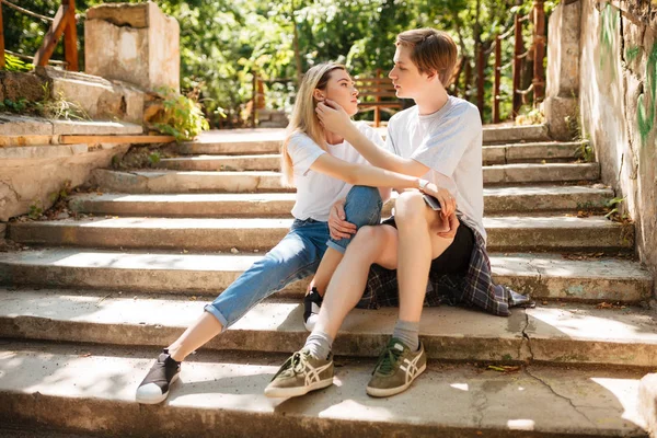 Giovane bella coppia seduta sulle scale nel parco e si abbracciano mentre trascorrono del tempo insieme. Ritratto di ragazzo e ragazza cool con i capelli biondi seduti e sognanti che si guardano l'un l'altro — Foto Stock