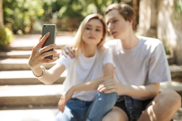 Porträt eines jungen schönen Paares, das sitzt und Selfies macht. Nahaufnahme Foto einer Frau, die ihr Handy in der Hand hält und ein Foto mit der Frontkamera des Handys macht — Stockfoto