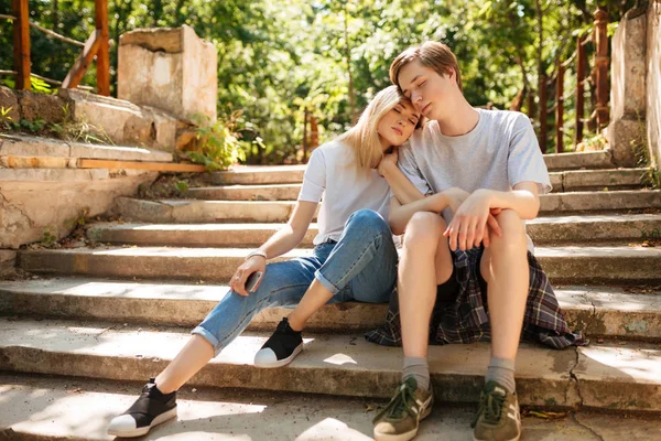 Mooie jonge koppel zittend op een trap in park en dromerig sluiten hun ogen terwijl het besteden van tijd samen. Portret van cool jongen en meisje met blond haar, leunend haar hoofd op zijn schouder in park — Stockfoto