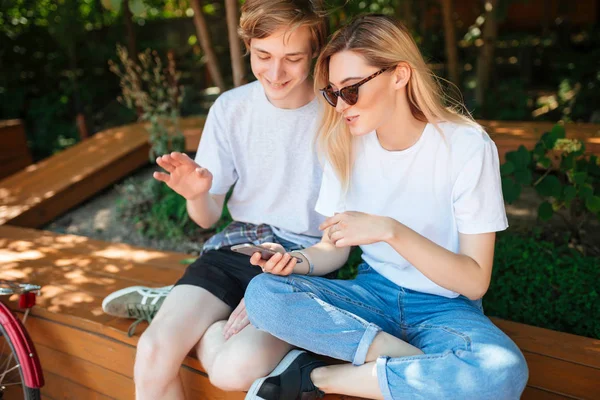 Κοντινό πλάνο φωτογραφία ζευγάρι κάθεται σε παγκάκι στο πάρκο και ευτυχώς συζήτηση κάτι κατά τη χρήση κινητών τηλεφώνων. Πορτρέτο του νεαρού και όμορφη κυρία με ξανθά μαλλιά ξοδεψουν το χρόνο μαζί — Φωτογραφία Αρχείου