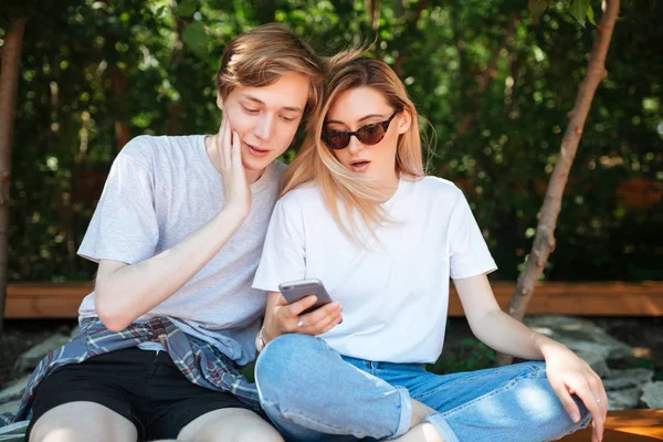 Πορτραίτο του καταπληκτικός ζευγάρι κάθεται στο παγκάκι στο πάρκο και περίσκεψη ψάχνει σε κινητό τηλέφωνο. Εσωτερικη φωτογραφία του νεαρού και ωραία κυρία με ξανθά μαλλιά που αφιερώνουν χρόνο μαζί, ενώ η χρήση κινητού τηλεφώνου — Φωτογραφία Αρχείου