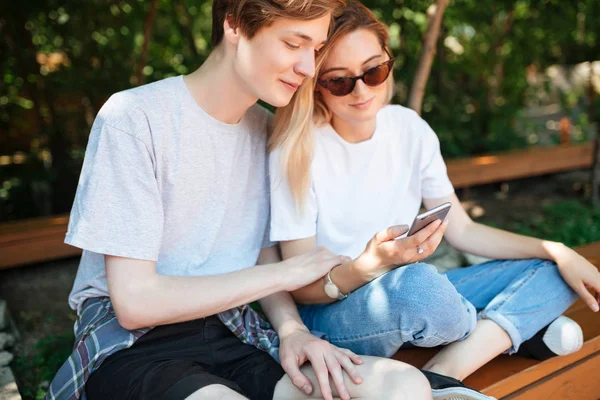 Parkta bankta oturmuş ve cep telefonu kullanarak güzel çift portresi. Fotoğraf güzel bayan ve genç adam kadar birlikte vakit elinde cep telefonu tutan sarı saçları ile kapatmak — Stok fotoğraf