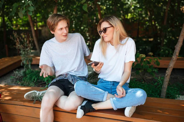 Πορτραίτο του ζευγάρι κάθεται σε παγκάκι στο πάρκο και συναισθηματικά συζήτηση κάτι κατά τη χρήση κινητών τηλεφώνων. Εσωτερικη φωτογραφία του καταπληκτικός άνθρωπος και όμορφη κοπέλα με τα ξανθά μαλλιά ξοδεψουν το χρόνο μαζί — Φωτογραφία Αρχείου