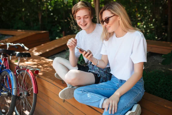 Glada par sitter på bänken i parken och glatt tittar i cellphone med två cyklar i närheten. Porträtt av ung leende man och vacker dam med blont hår spendera tid tillsammans — Stockfoto