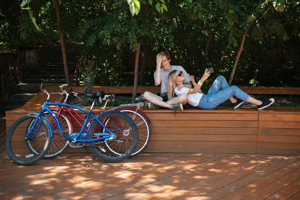 Młoda para zabawy w parku z czerwonego i niebieskiego rowerów w pobliżu. Fajny chłopak siedząc na ławce w parku z piękną dziewczyną z blond włosy że opierając się na niego i biorąc selfie razem na telefon — Zdjęcie stockowe