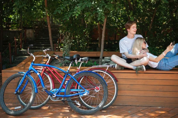 Foto de cerca de las bicicletas rojas y azules en el parque. Niño sentado en el banco en el parque con chica bonita con el pelo rubio que se apoya en él y tomar selfie juntos en el teléfono celular —  Fotos de Stock