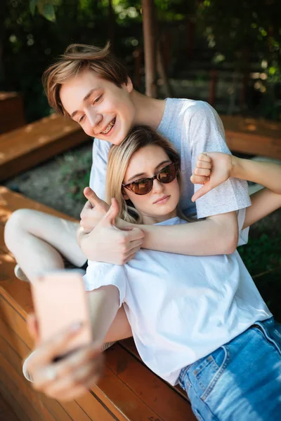 Porträtt av pojke sitter på bänken i parken och lyckligt visar tummen medan tjej med blont hår lutande på honom och visar tummen ner. Unga par med söt selfie samtidigt spendera tid tillsammans — Stockfoto