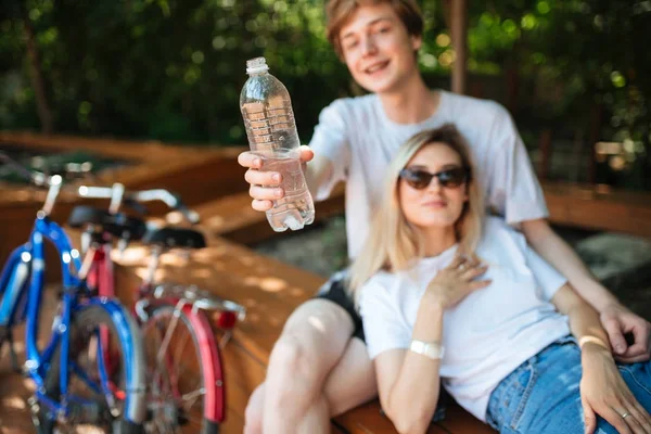 Närbild foto av mannen handen håller flaska vatten i park med två cyklar i närheten. Ungt par sitter på bänken och glatt tittar i kameran medan spendera tid tillsammans — Stockfoto