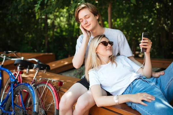 Ung cool par i hörlurar lyssna musik i parken med två cyklar i närheten. Porträtt av pojke sitter på bänken och drömmande stänga ögonen medan flicka lutande på honom med mobiltelefon i handen — Stockfoto
