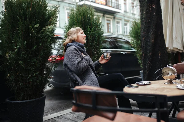 Portrait of joyful lady with blond hair sitting with cup of coffee in hand and happily looking aside on street isolated