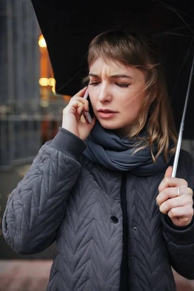 Portrait of upset lady standing on street with black umbrella and talking on her cellphone  isolated