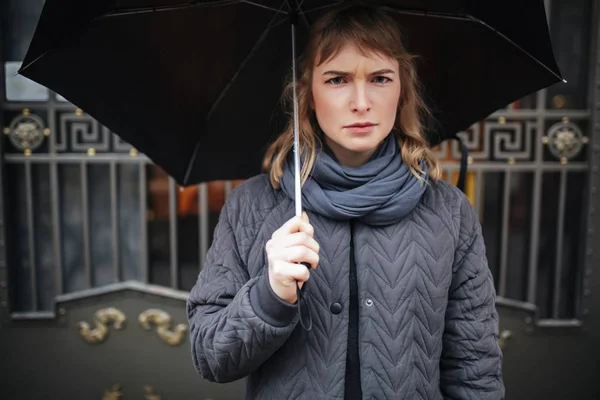 Portrait of upset lady standing on street with black umbrella and  sadly looking in camera isolated