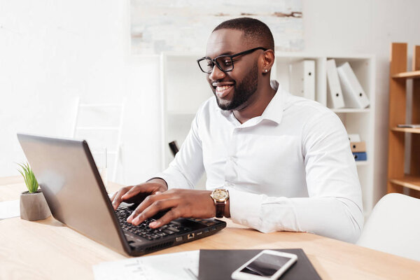 Portrait of smiling african american businessman in white shirt and eyewear sitting and working on his laptop in office isolated