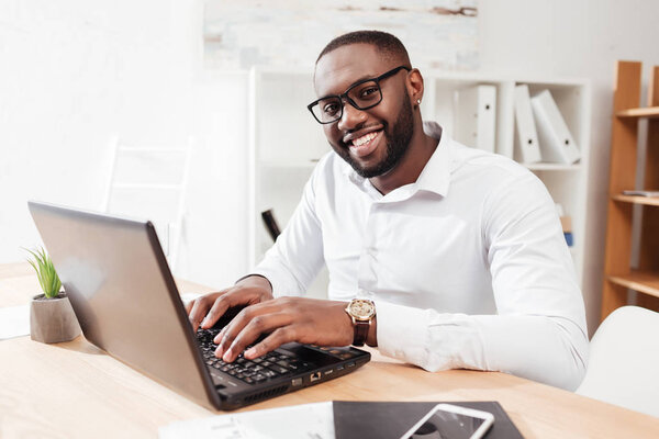 Портрет улыбающегося африканского бизнесмена в белой рубашке и очках, сидящего и счастливо смотрящего в камеру во время работы над ноутбуком в офисе изолированного
