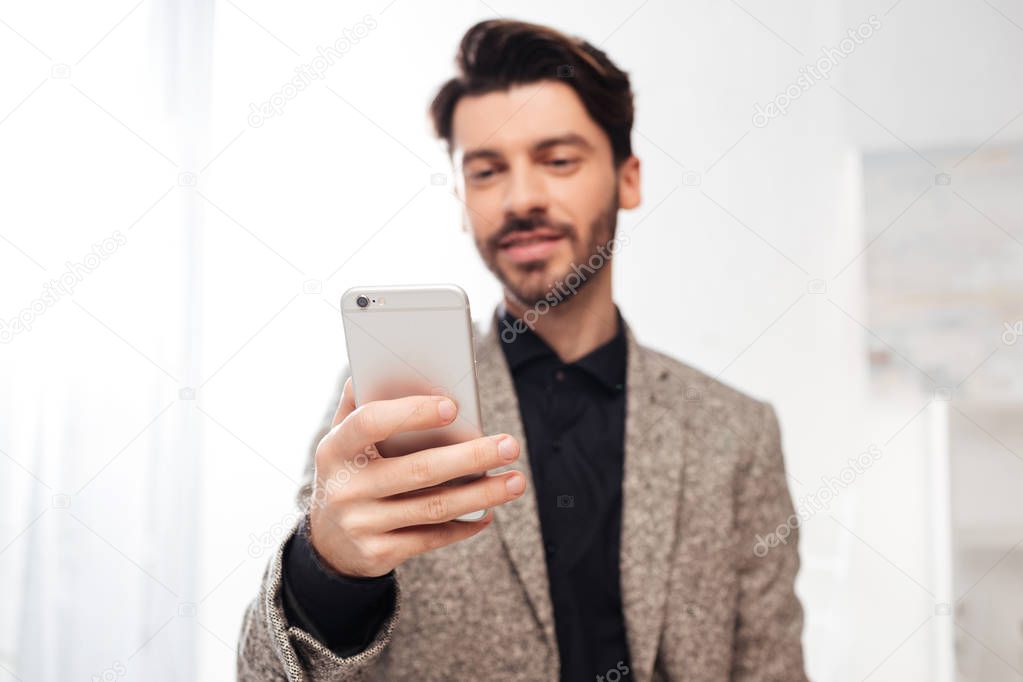 Close up photo of young businessman in black shirt and jaket standing and holding smartphone in hand in office isolated