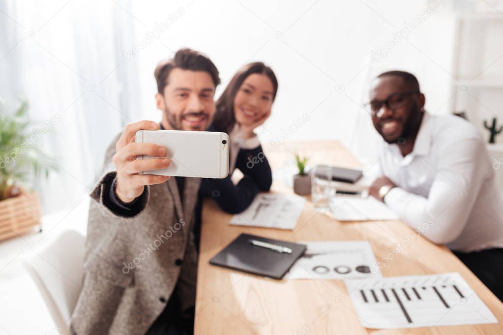 Portrait of multinational businessmen and businesswoman sitting at the table and taking photos on cellphone while working together in office isolated