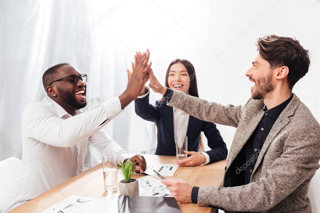 Portrait of smiling multinational group of business partners sitting at the table and happily giving high five to each other while working together in office isolated