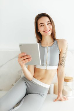 Sportif üst ve el ve sevinçle kamera izole arıyor tablet ile mutfak masasında oturan tozluk gülümseyen güzel Bayan