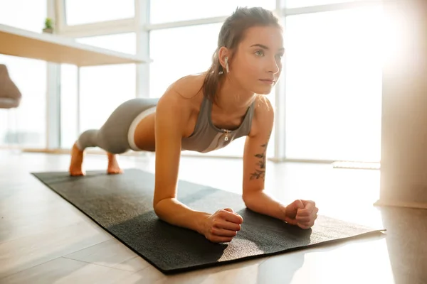 Sportif üst ve fitness yoga mat üzerinde pratik ve arka plan üzerinde büyük camları olan evde kulaklık müzik dinleme tozluk güzel Bayan portresi